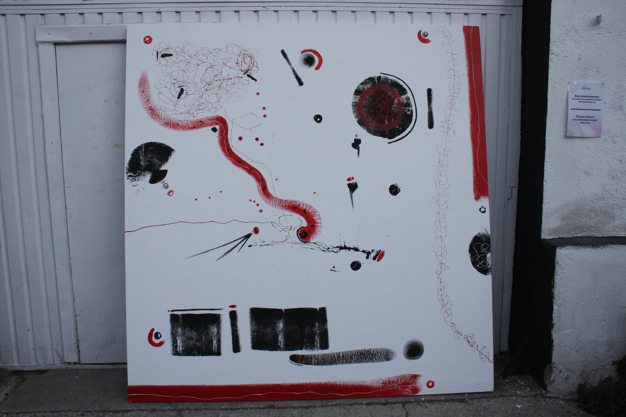 A large canvas leaning against a white wall outdoors, showing black bars, red marks and a red band along the bottom