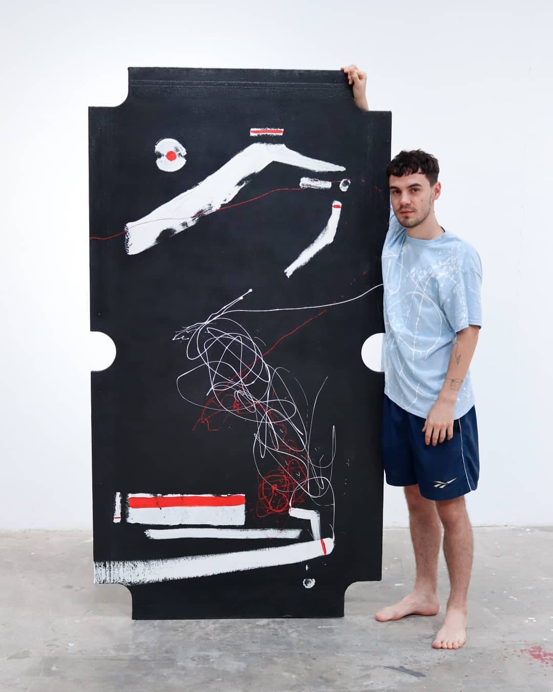 Barefoot in the studio, holding the tall notched black panel upright against a white wall