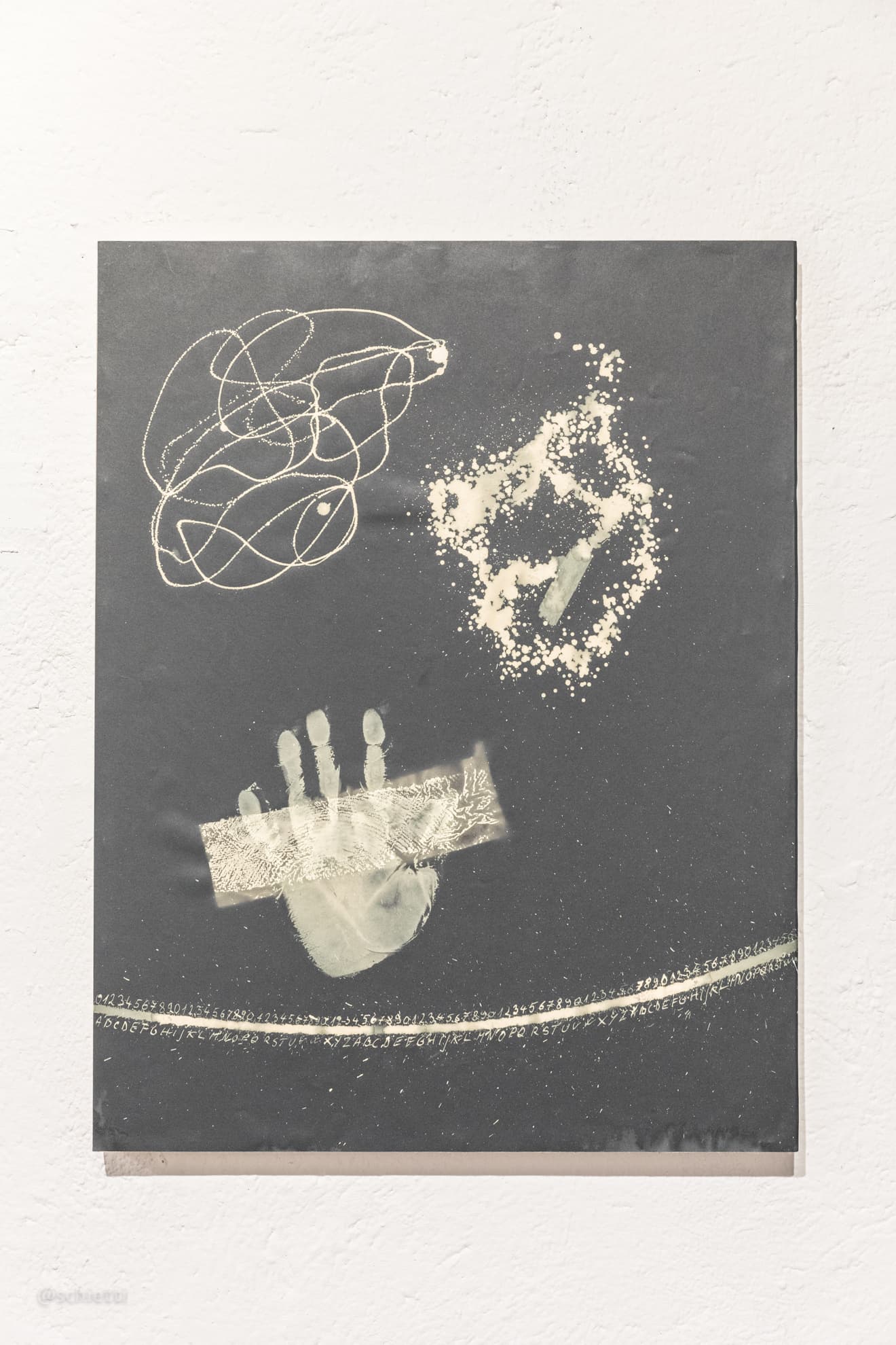 The whole sheet framed under glass on a white wall: a looping white scribble top left, a speckled burst top right, the bleached handprint in the middle and a long bright curve of small marks across the bottom