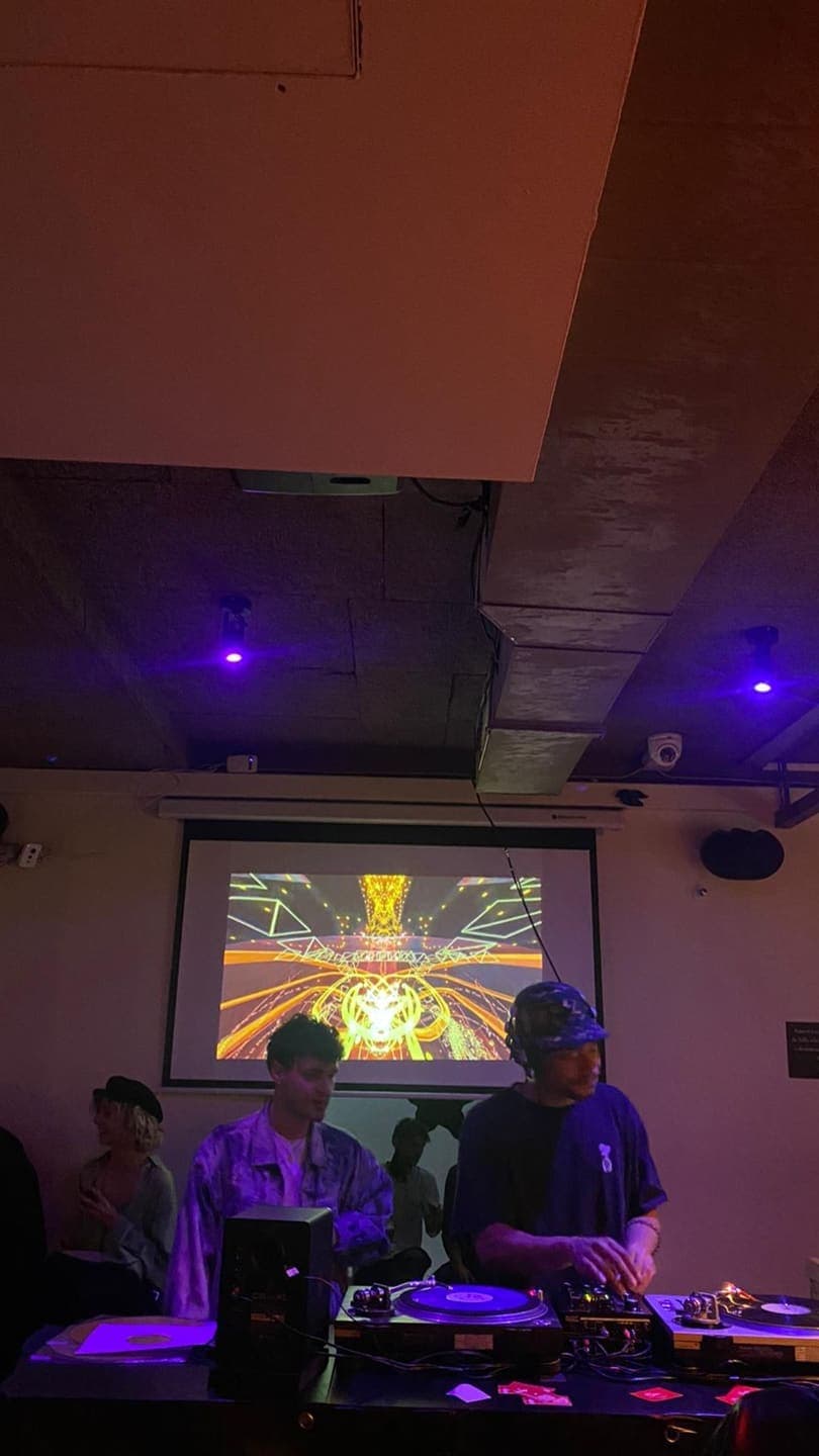 A dark club room with a projection screen above two DJs, showing a gold and orange symmetrical burst of light
