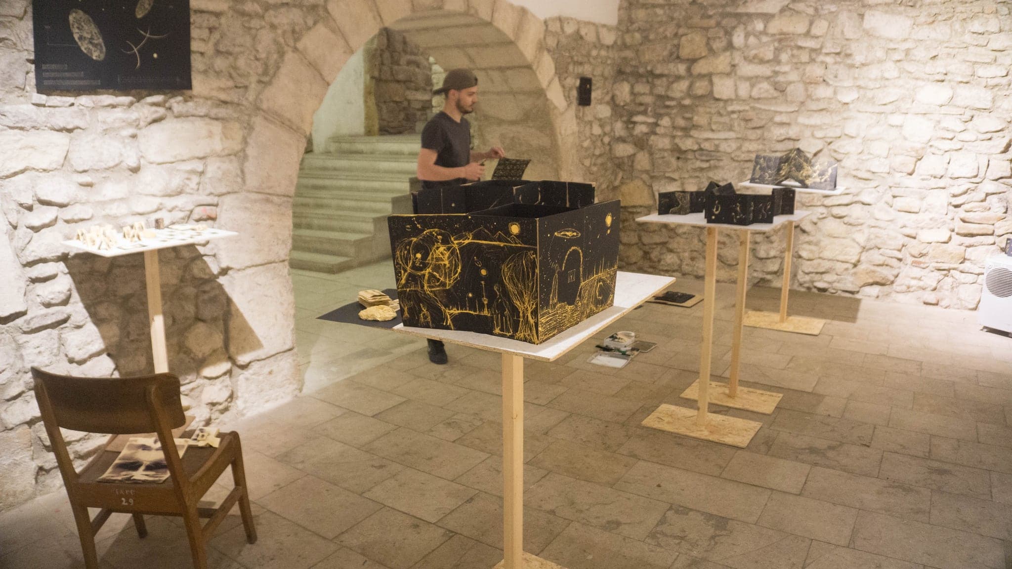 A stone-vaulted gallery with black and gold folding works standing open on pale wooden plinths, the artist working at one of them under the arches