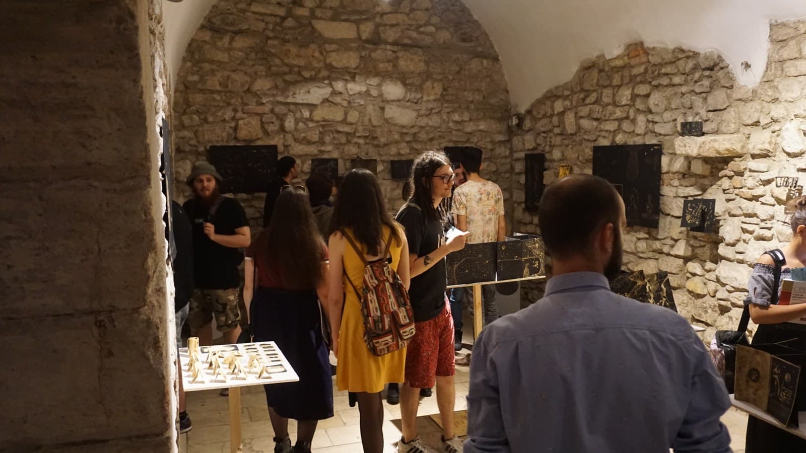 The vaulted stone cellar full of people at the opening, black-and-gold bleach panels standing on plinths along the wall and a table of small drawings in the foreground