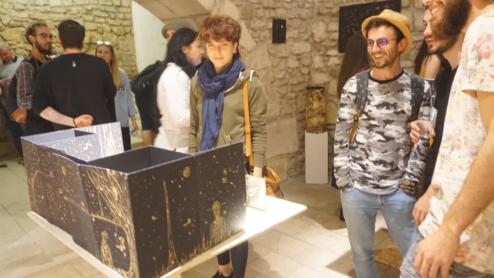 A large folding black panel covered in pale gold marks standing open on a plinth, guests talking around it in the stone room