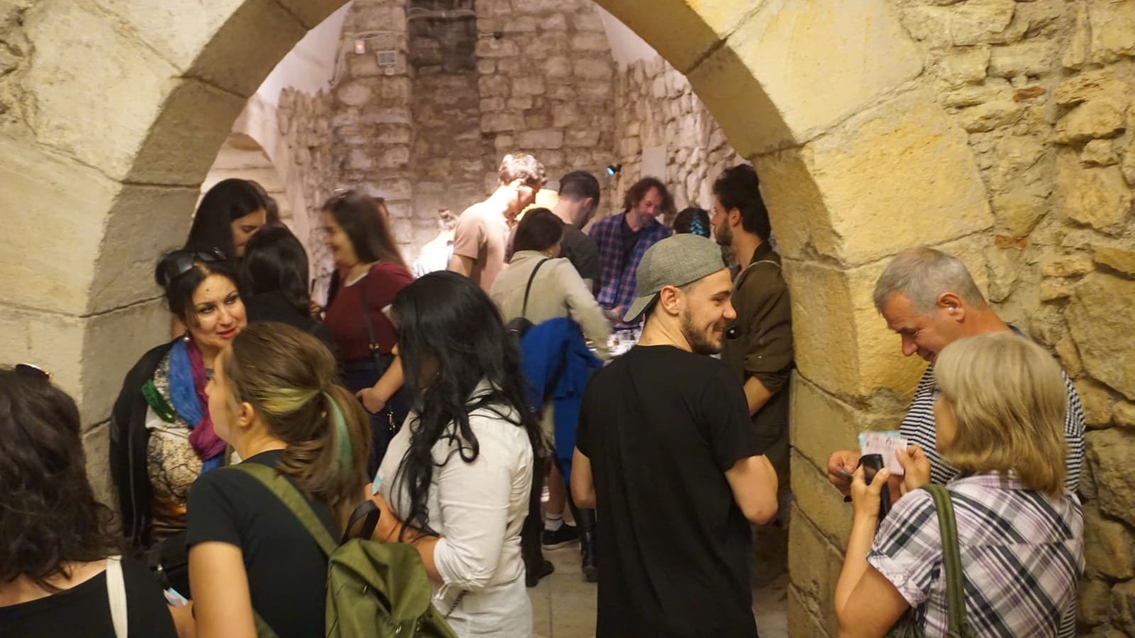 A crowd gathered under a tall stone arch at the entrance in warm light, one person raising a camera