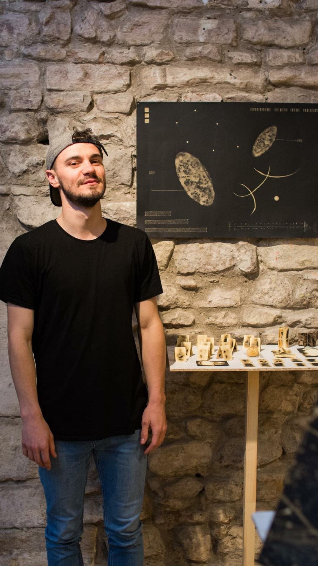 Ovidiu standing against the stone wall of Galeria Casa Matei beside a large black panel of pale ovals and dotted lines, with wax works laid out on a table to his right