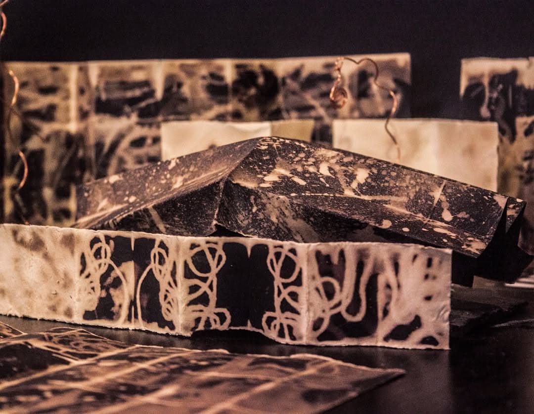 Folded wax and paper sheets standing on a dark table, marked with black chemical blooms and looping pale lines, more sheets ranged behind them