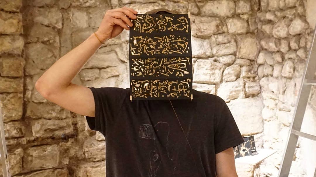 A folding book of black panels covered in pale gold marks, held up in front of the face against a stone wall