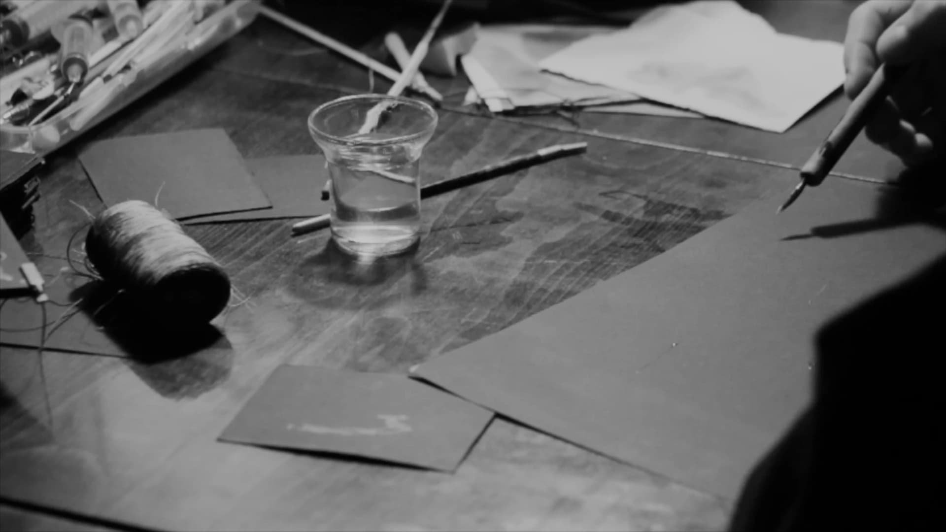 Black and white view of a work table, a glass of clear liquid, a dip pen held in one hand, sheets of black paper, a spool of thread and a box of tools