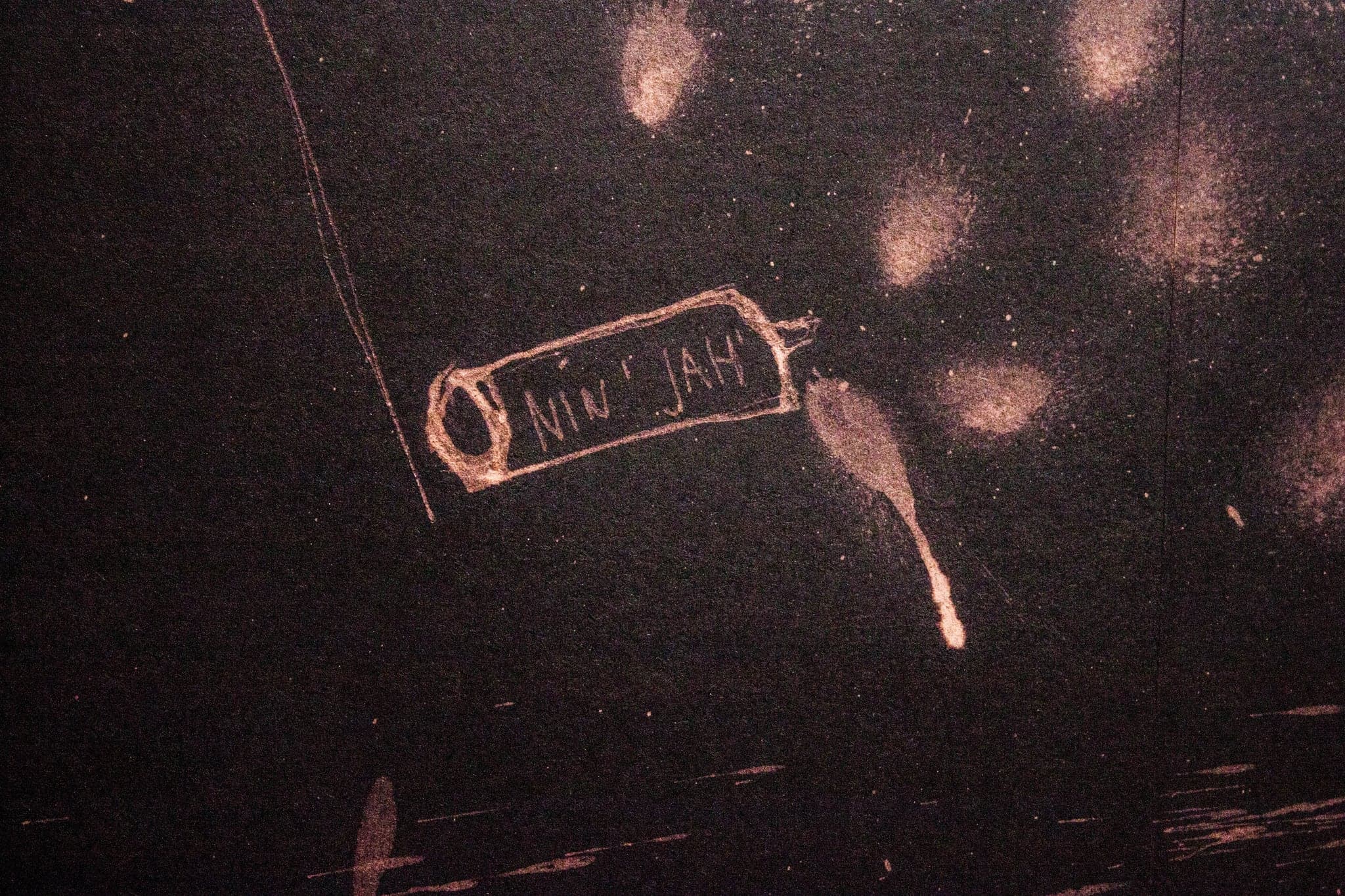 Close photograph of black paper showing its grain, with a small pale cartouche drawn in bleach reading NIN JAH, a run drip below it and soft bleached blooms to the right.