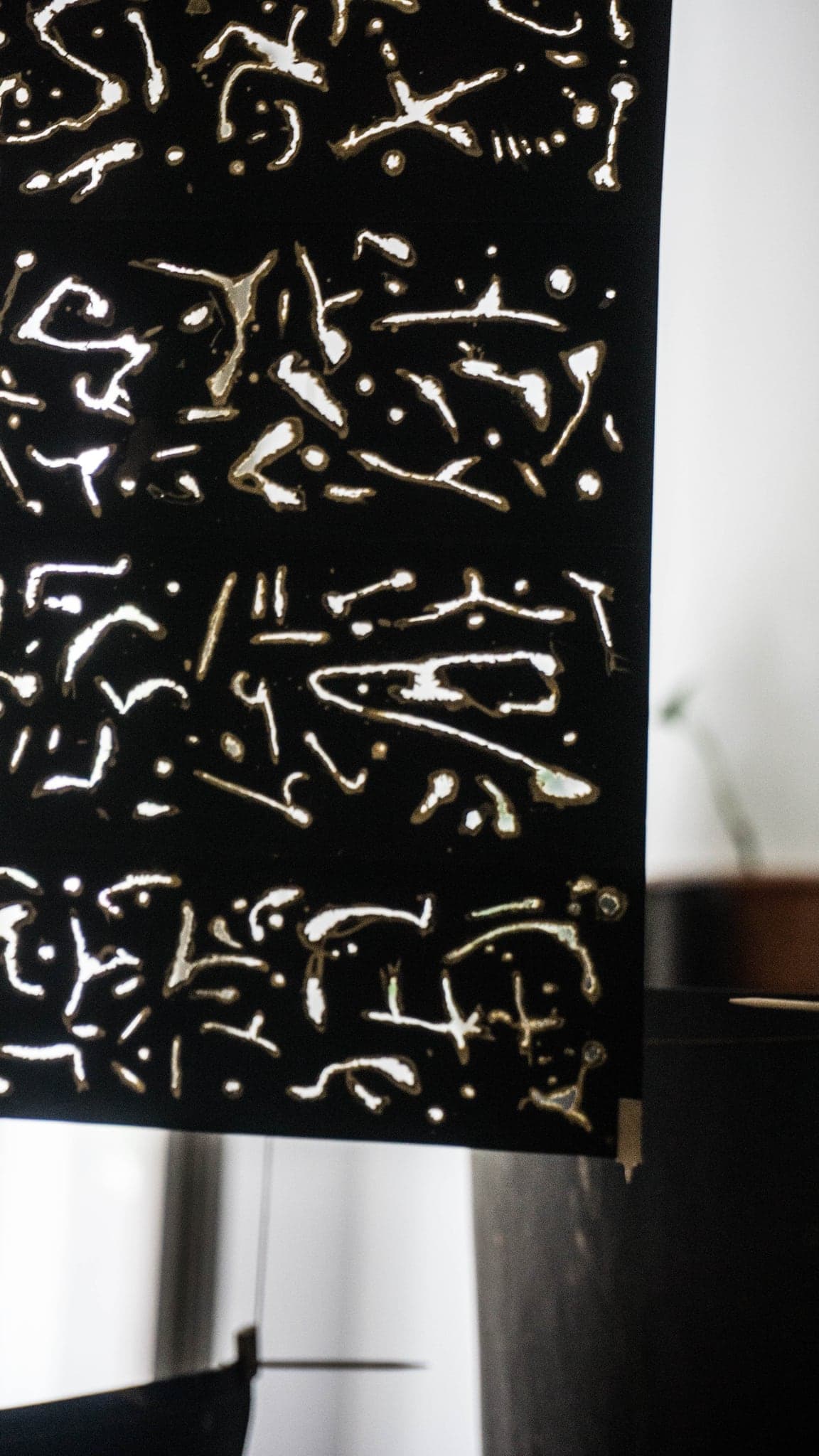 A black sheet of paper hung from a rail in a daylit room, covered edge to edge with pale glyph-like marks.