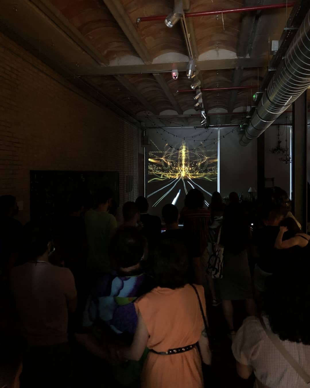 A room of people standing in the dark with their backs to the camera, watching the projection at the end of a brick hall