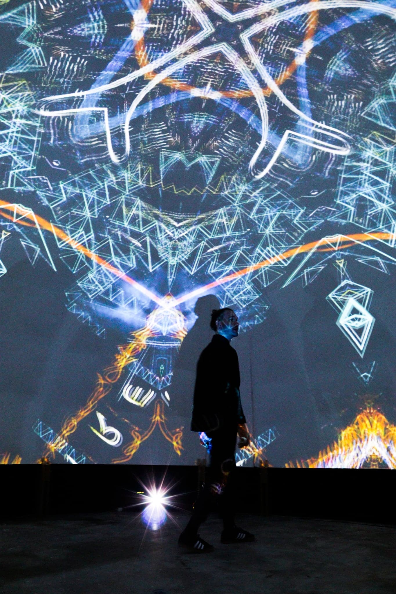 A visitor in silhouette walking across the dome floor, the projector's flare at their feet and a pale mandala filling the shell behind them