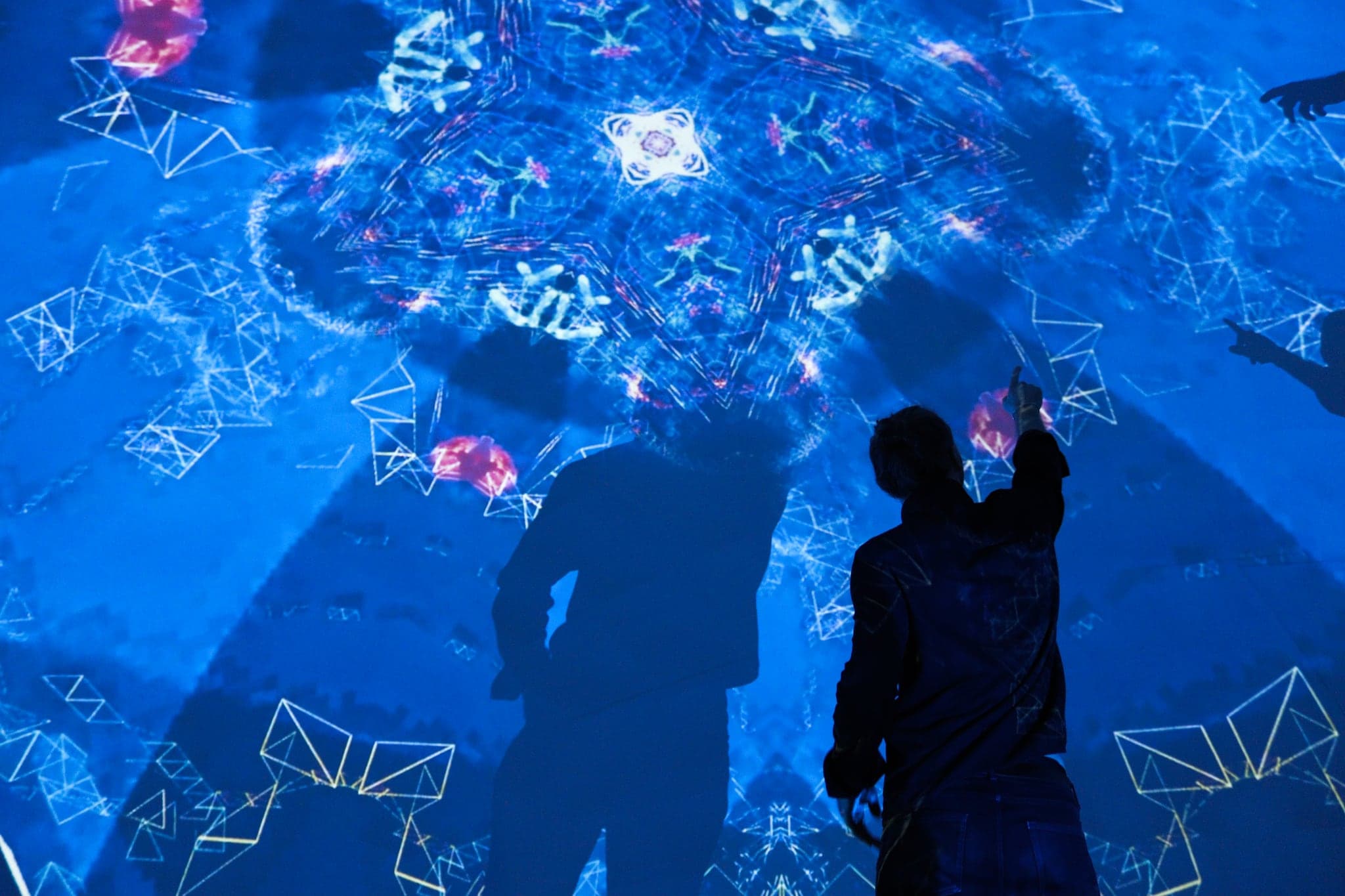 Two people seen from behind in deep blue light, one pointing up at a ring of drawn flowers and wire triangles overhead