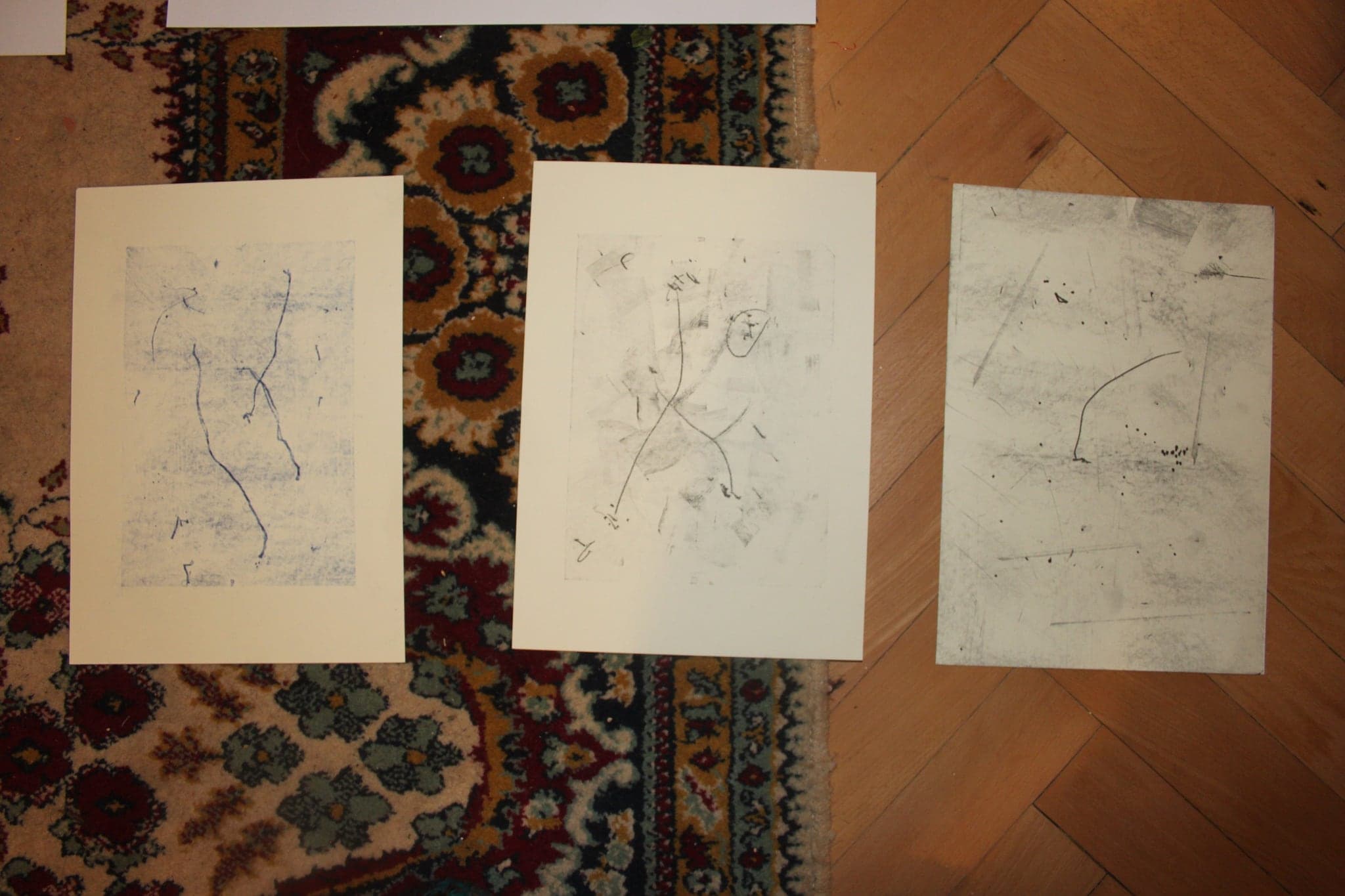 Three large pale drawings laid side by side on a patterned carpet, each with fine graphite lines and small dark forms