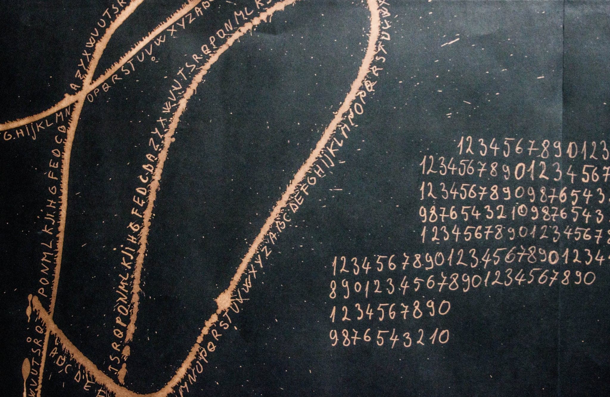 Alphabets written in pale gold along long curving lines, with columns of handwritten digits counting up and back down at the right