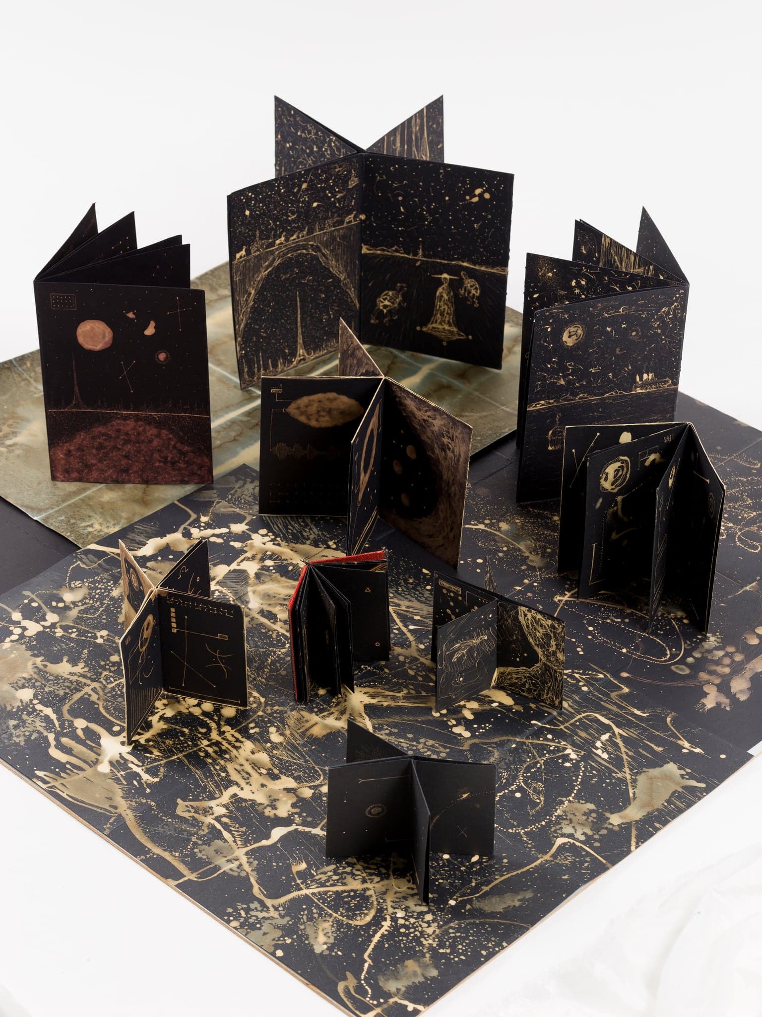 About ten small black concertina books standing open together on a large pale sheet that has been bleached into a splattered star map, on a white ground.