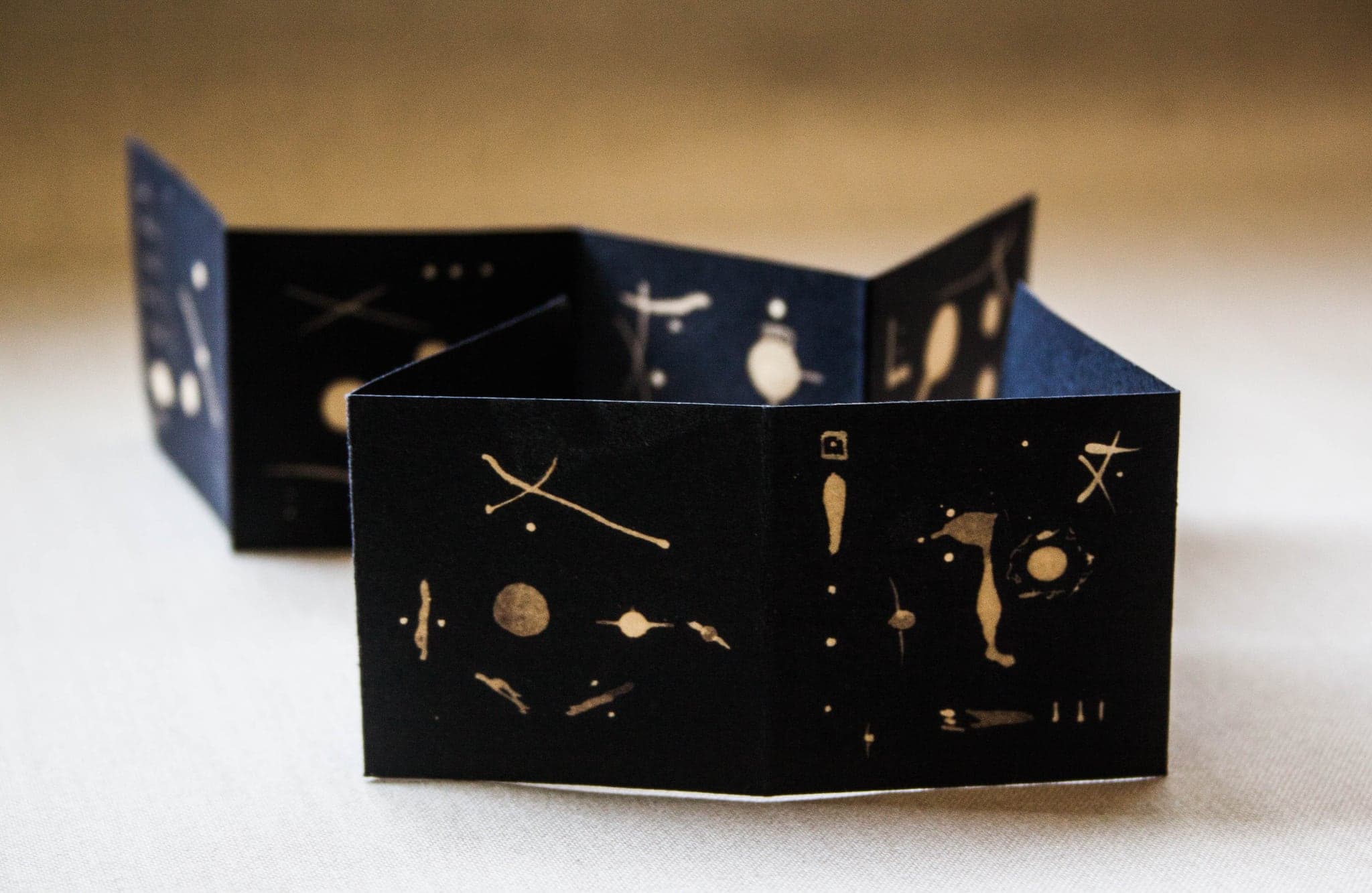 A small black concertina book standing open on a cream cloth, its panels marked with pale dots, crosses and a round moon shape.
