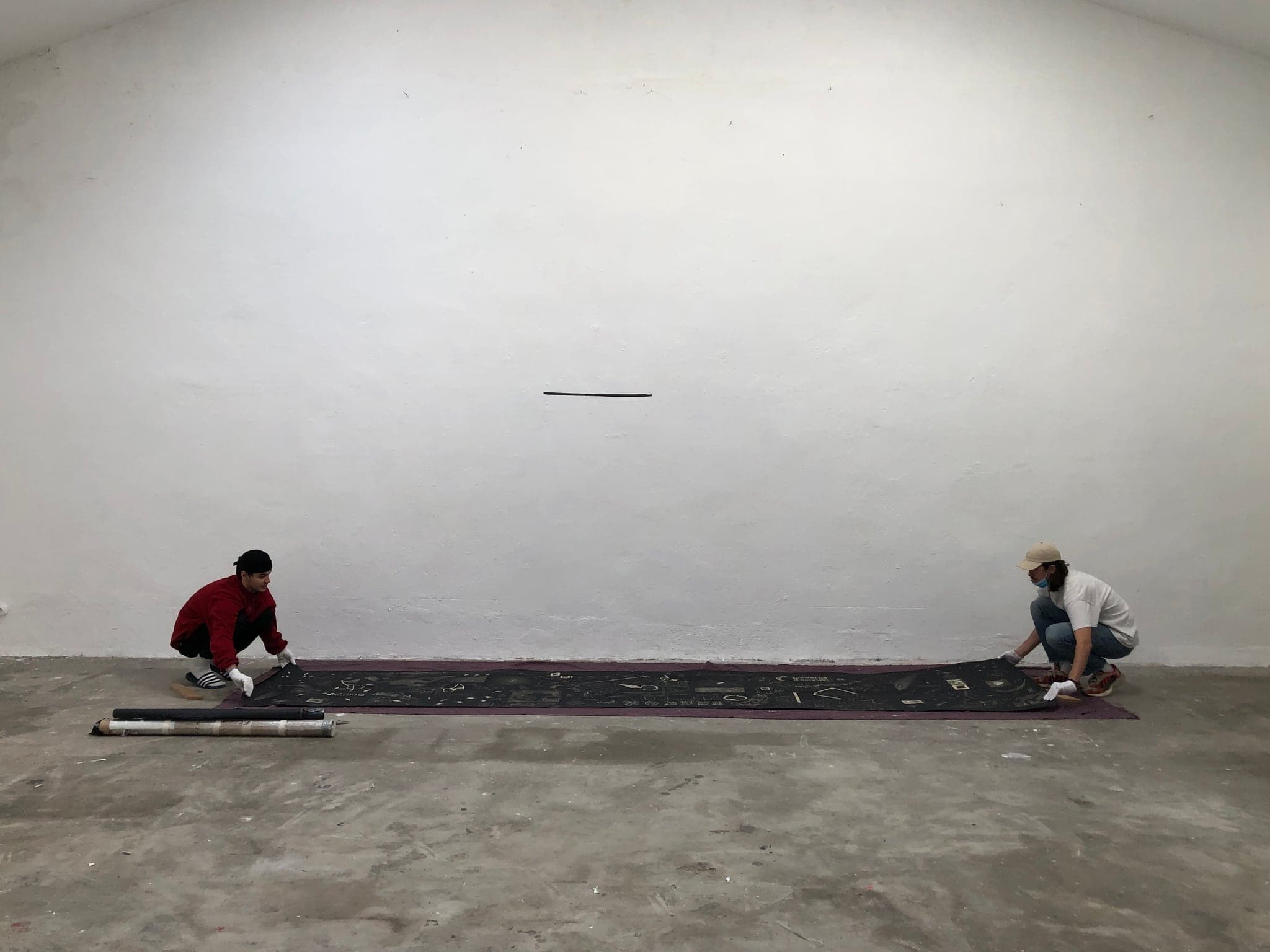 The full length of the black panel unrolled flat across a concrete gallery floor, two people crouched at either end, a spirit level lying beside it