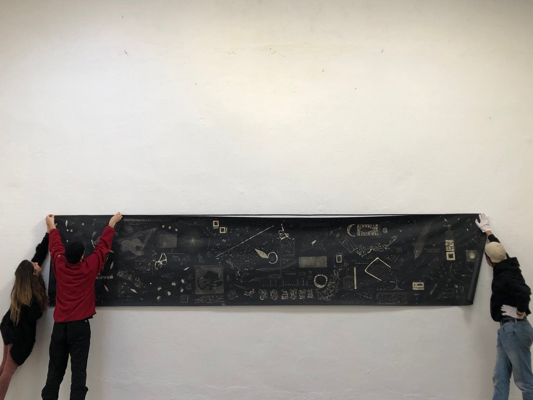 Three people holding the whole five-metre panel up flat against a white wall at head height, its bleached figures visible along its length