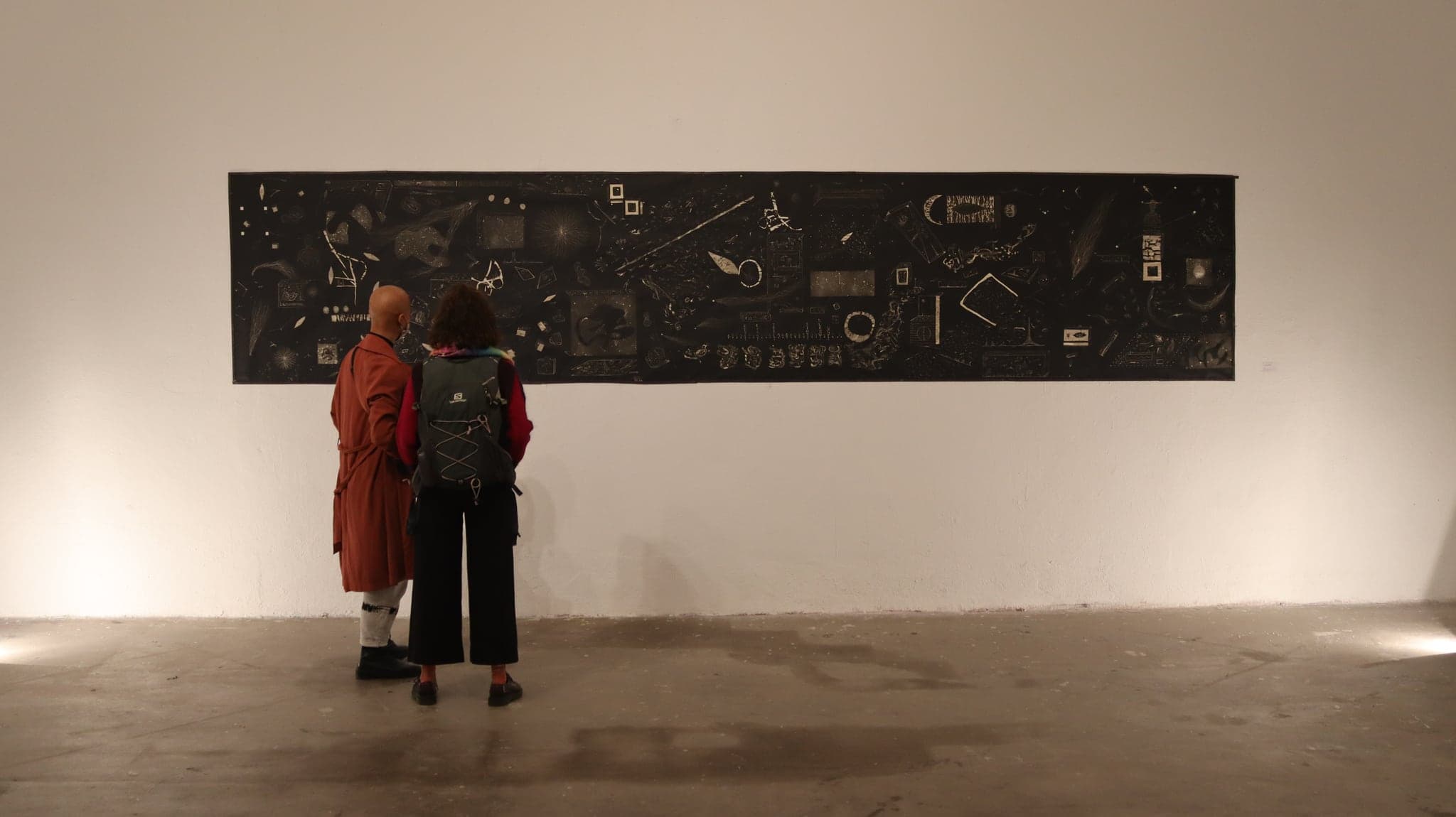 The panel hung and lit on a white gallery wall, a visitor in a red coat standing beneath it for scale in a large empty room