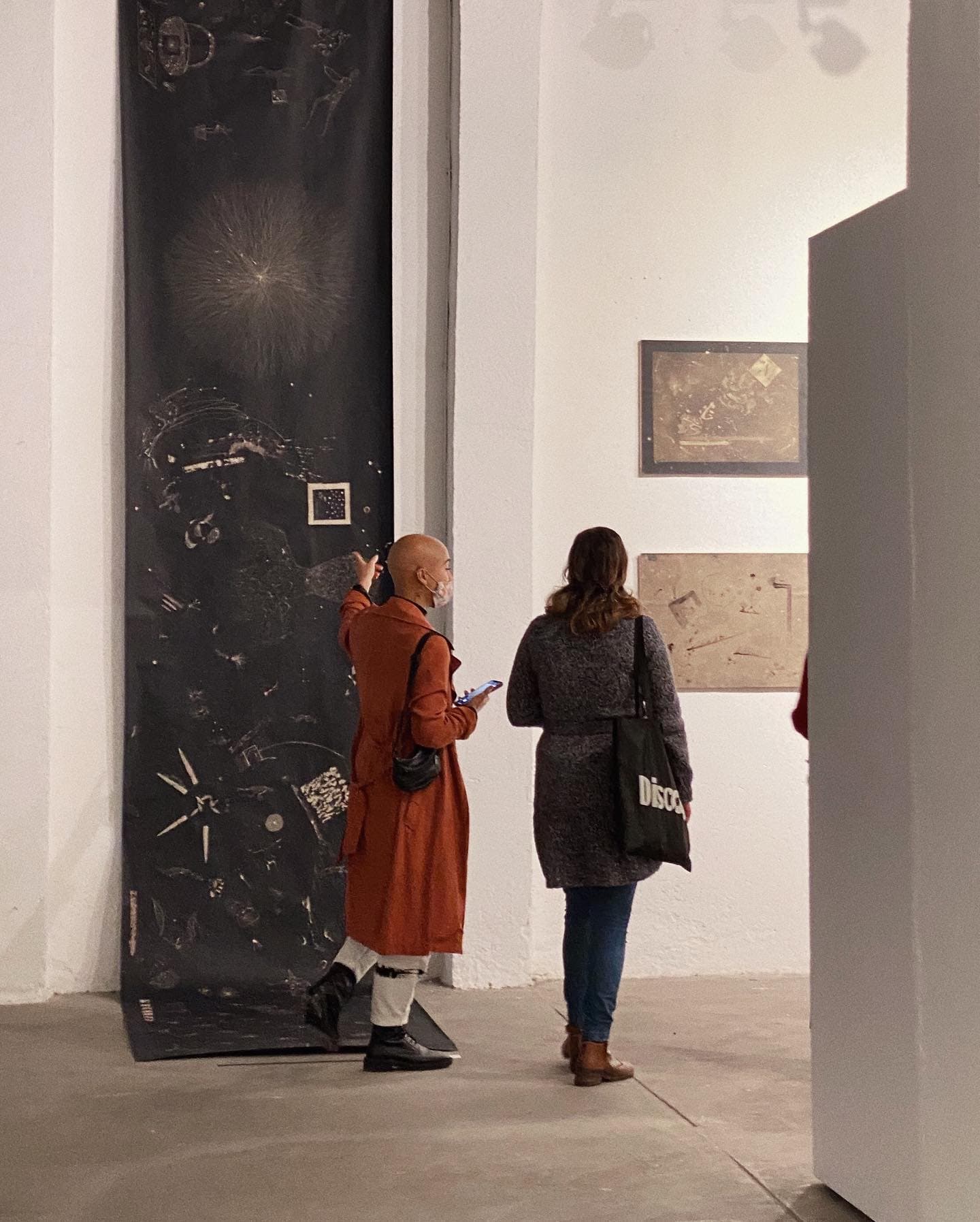 The same panel hung vertically from the ceiling, its lower end rolling out onto the concrete floor, with a visitor in a red coat reaching up to point at one of the figures