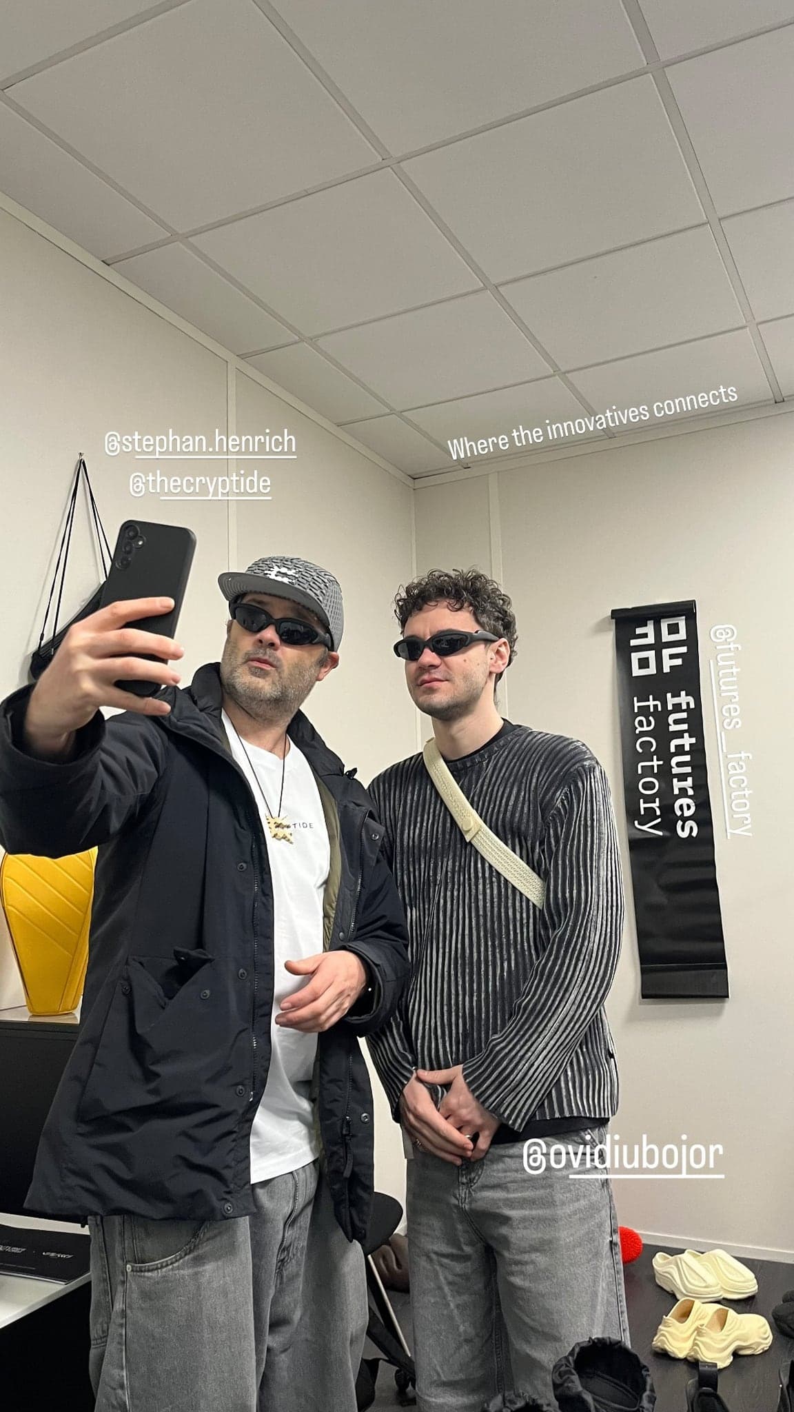 Two designers taking a phone selfie in the Futures Factory booth, black banner on the wall behind them
