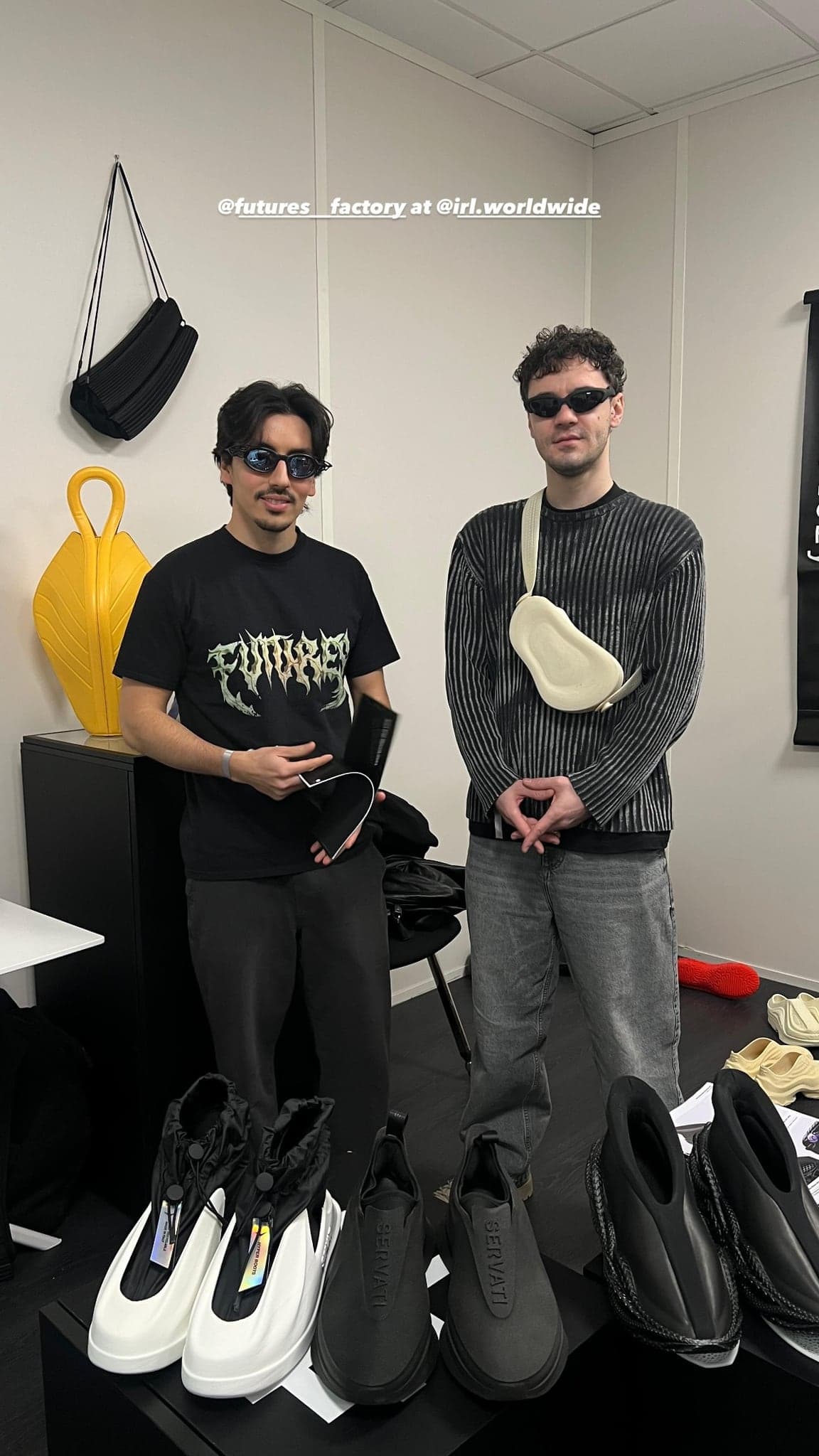 Two people standing behind a black plinth of sculptural shoes in a small white showroom, a yellow moulded vase on the cabinet behind