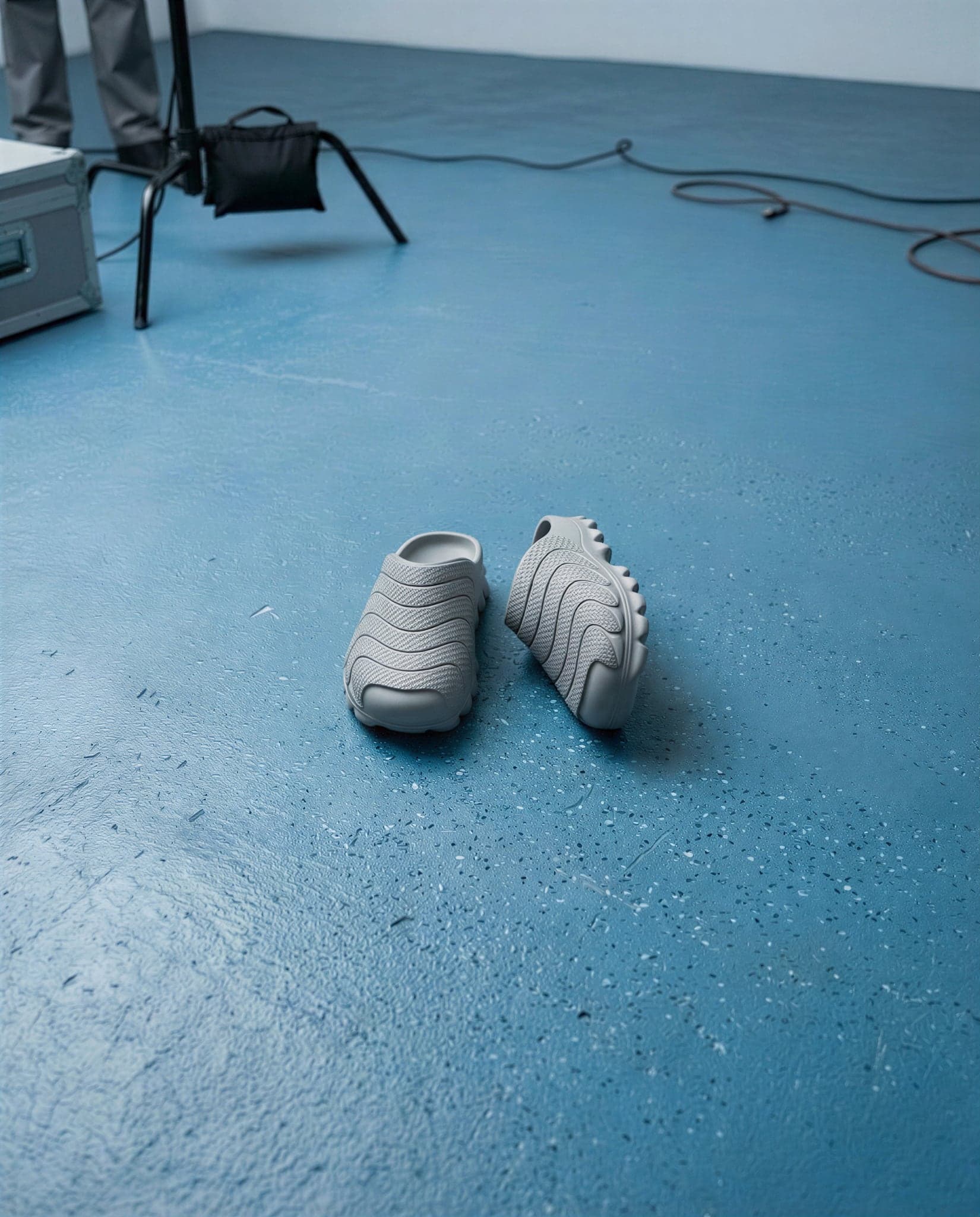 The pair of mules left on a wide empty blue studio floor, one tipped onto its side showing the lugged outsole.