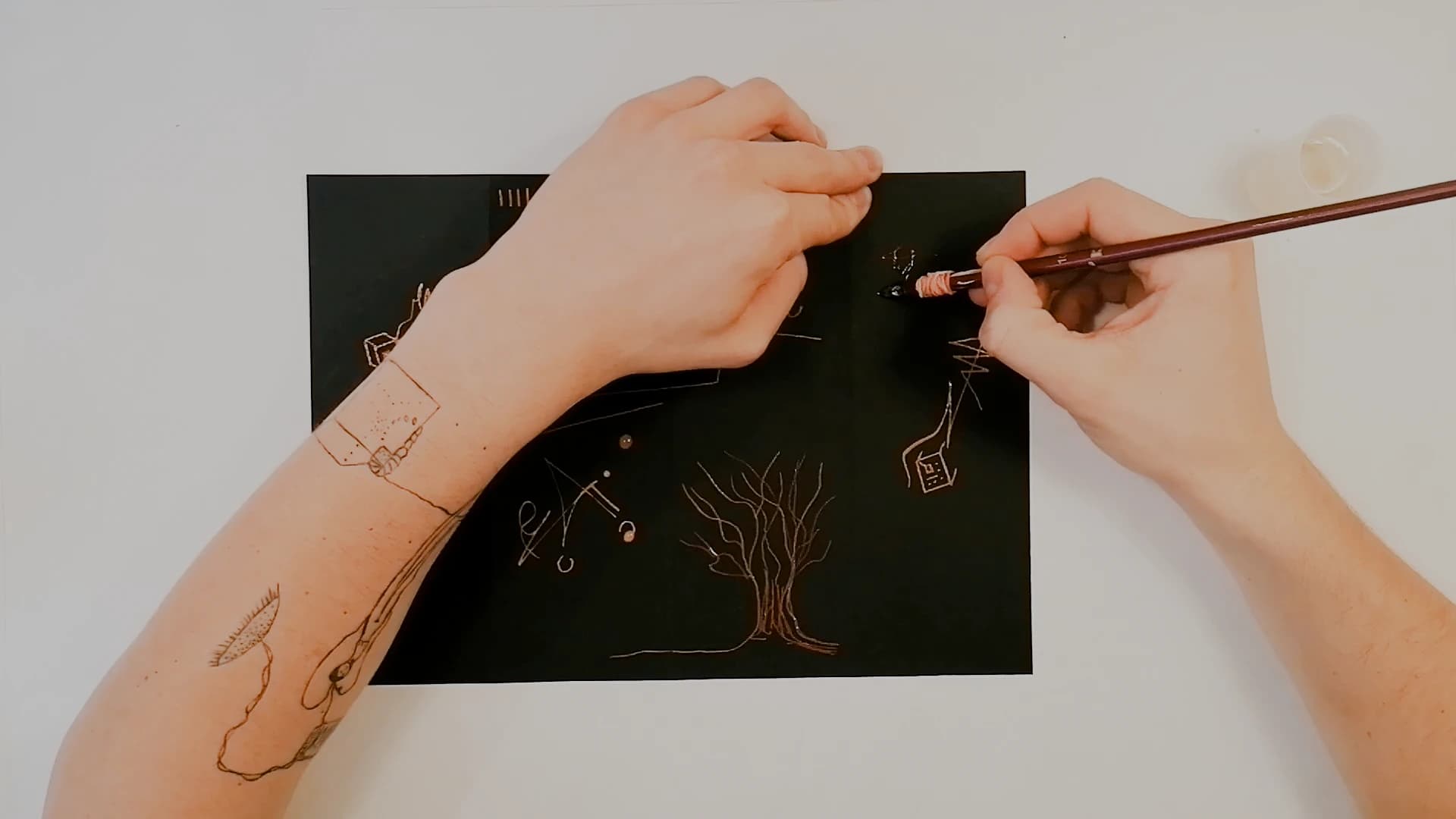 The black sheet part-filled. A bare tree with fine branches has been drawn in pale gold; a forearm crosses the frame, its tattoo echoing the marks on the paper.