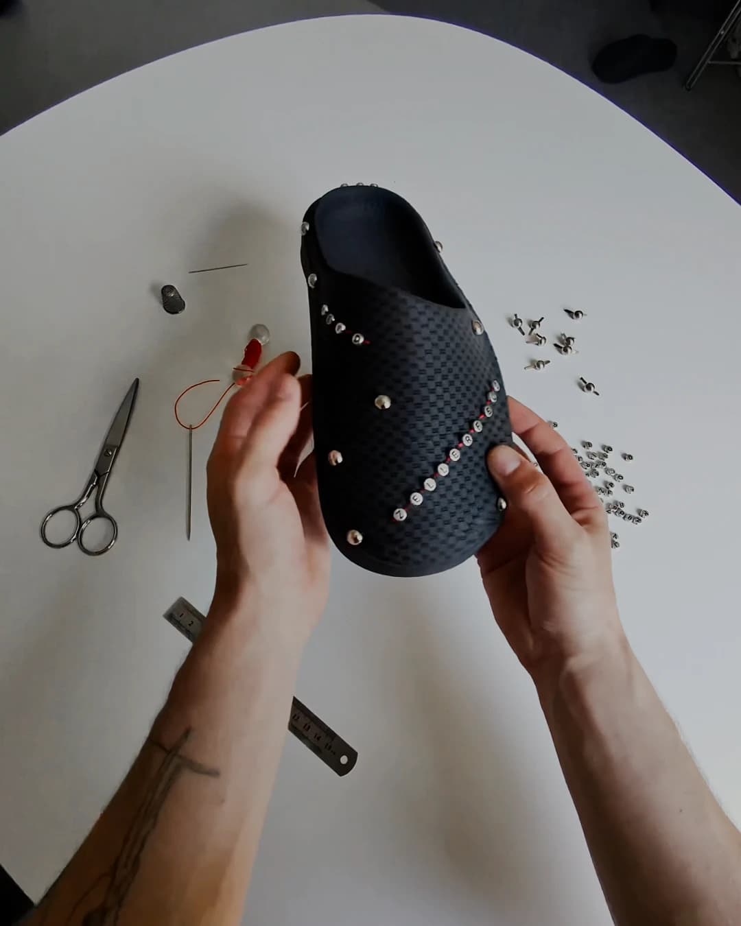 Overhead view of the black shoe held in two hands on a white table, scissors and a ruler beside it, the first studs scattered across the surface