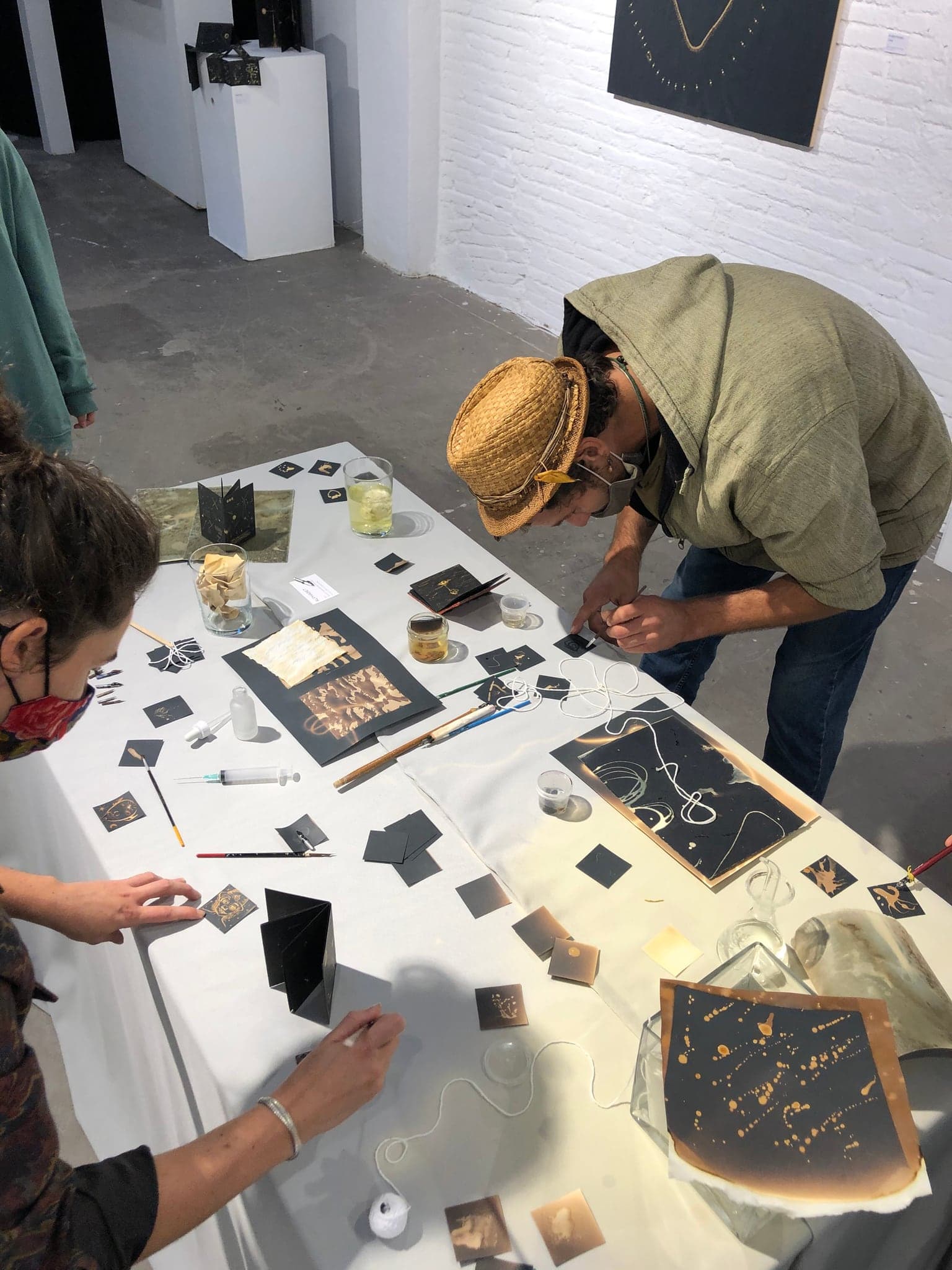 Two visitors bent over the white table working on black cards with brushes, glasses of bleach and finished sheets around them