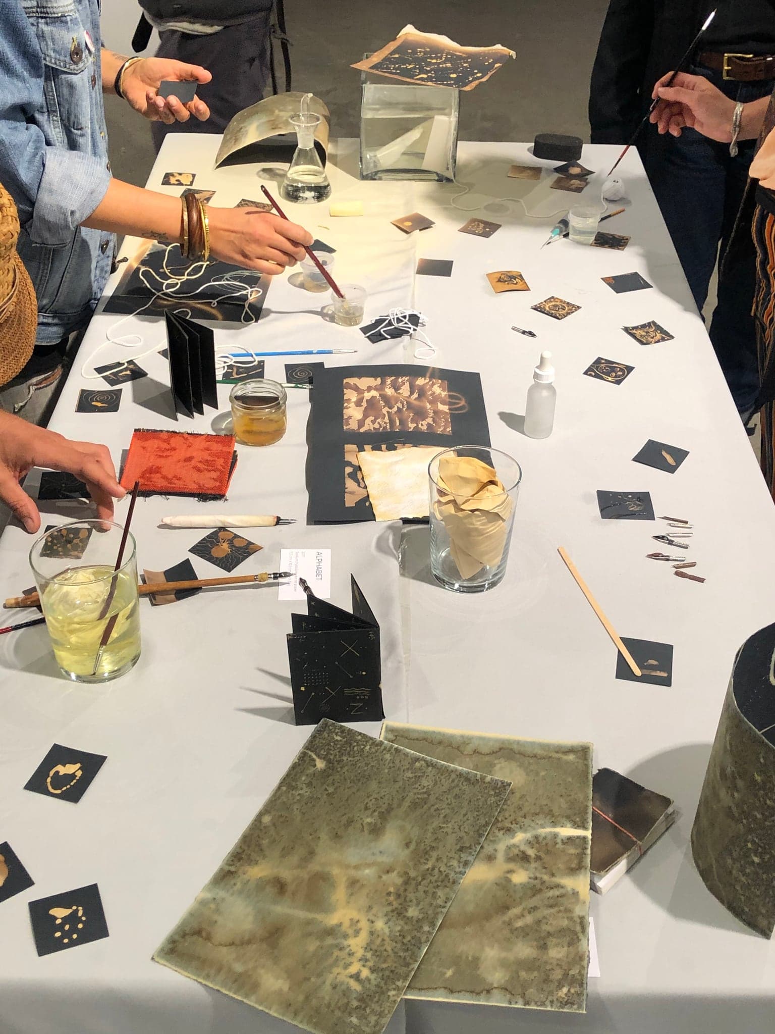 Several pairs of hands over the table at once, brushes and droppers working on small black cards, a large gold-marked sheet in the middle