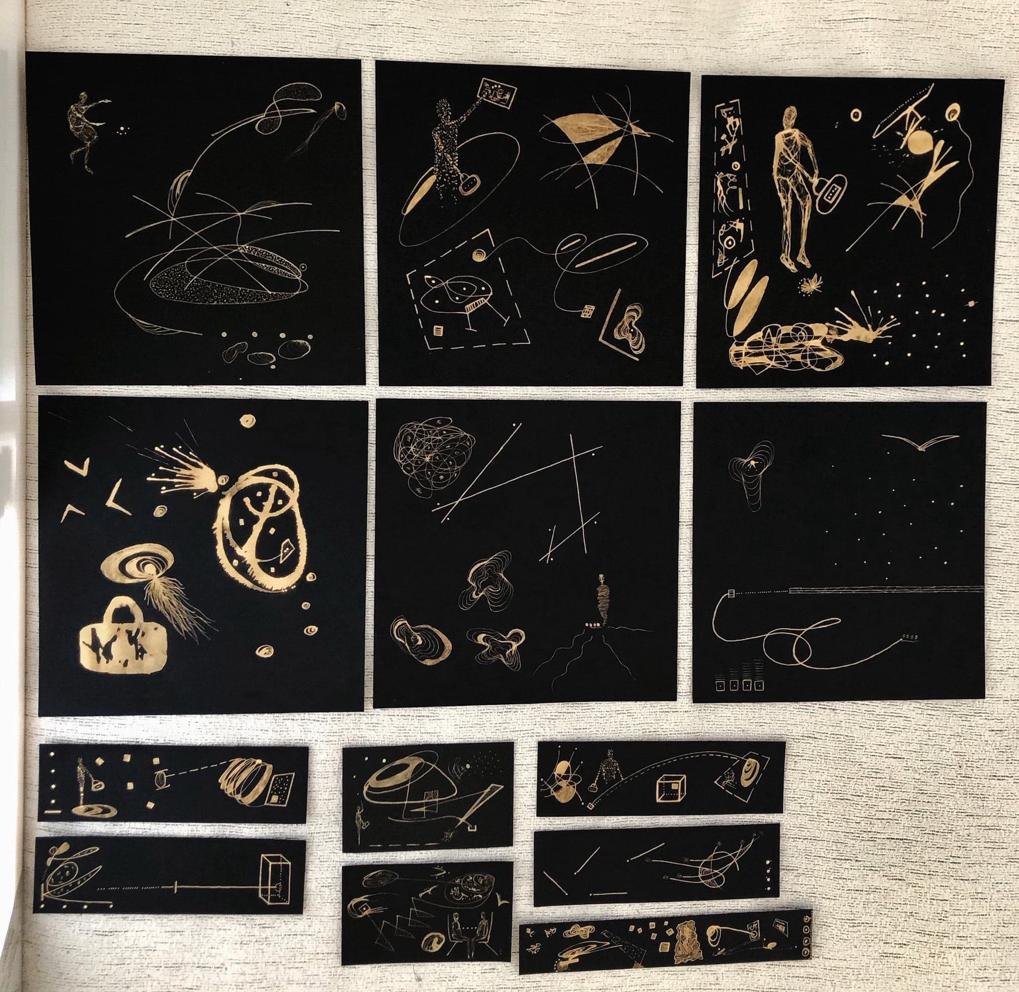 A group of black works with gold drawing laid out on a woven mat, six squares above, four long horizontal strips below