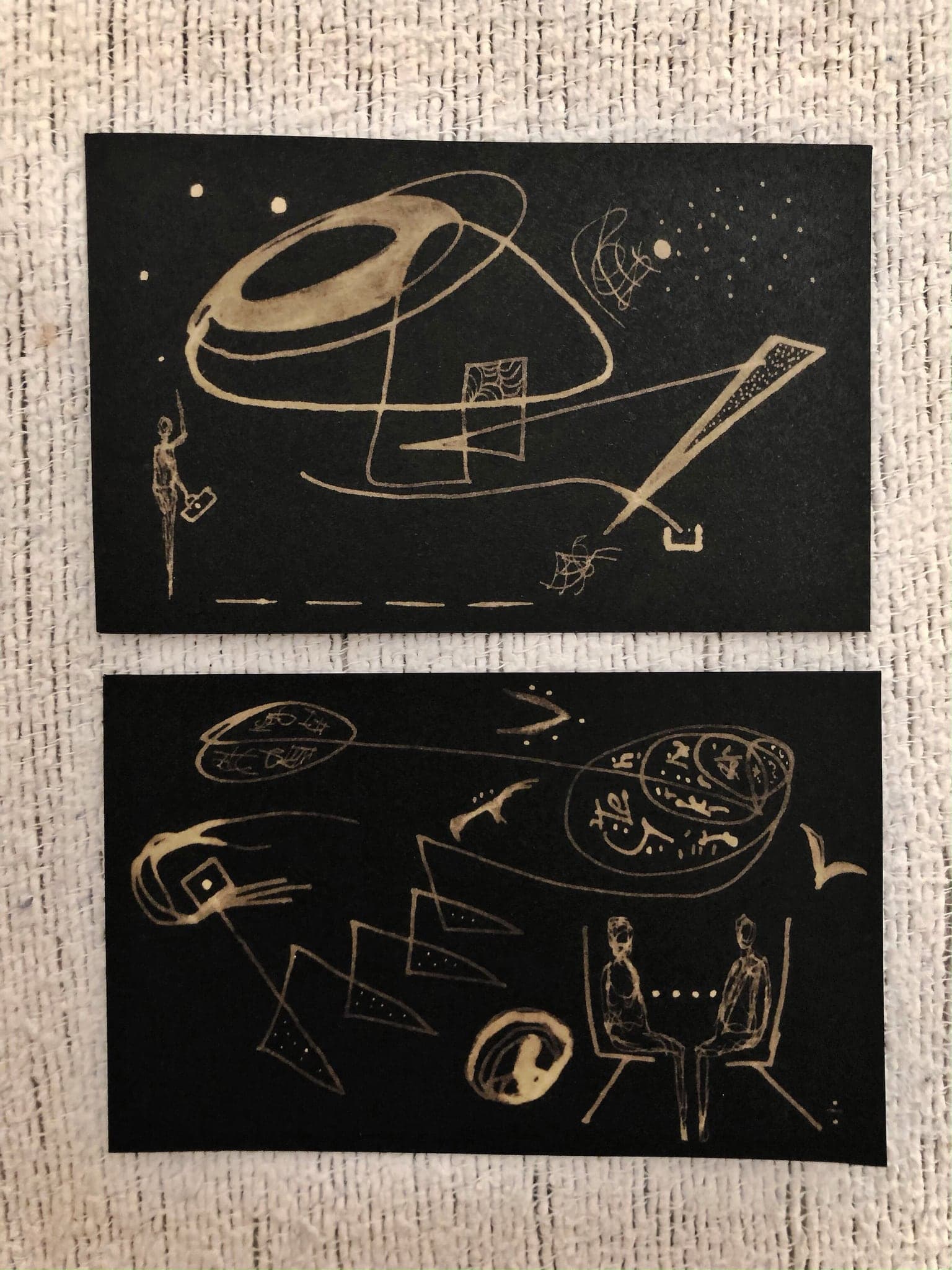 Two black works photographed on a woven mat, one with a large pale ring and a long ruled bar, the other with seated figures and small framed signs