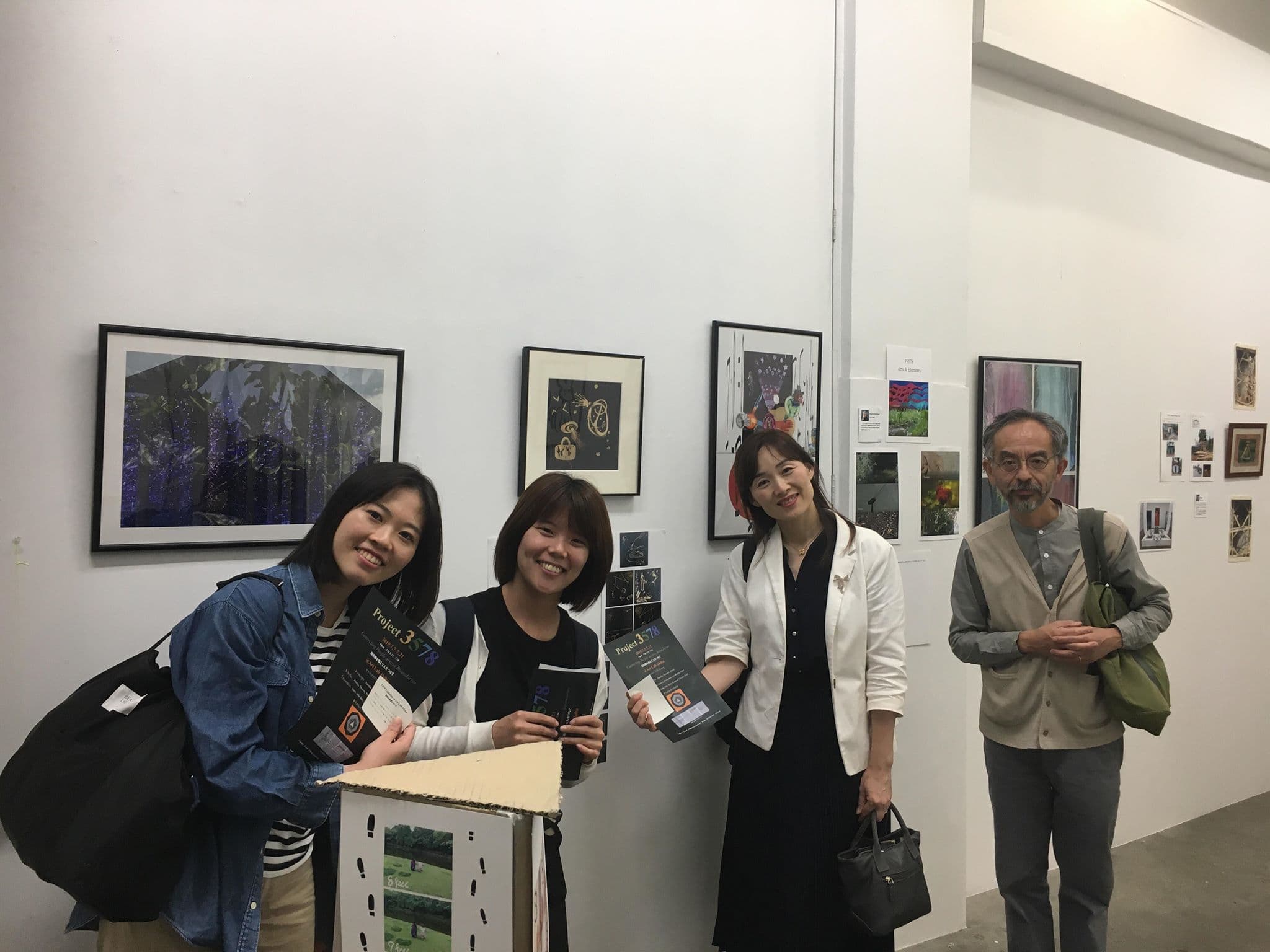 Four visitors standing in the gallery in front of a wall of framed works, holding printed programmes