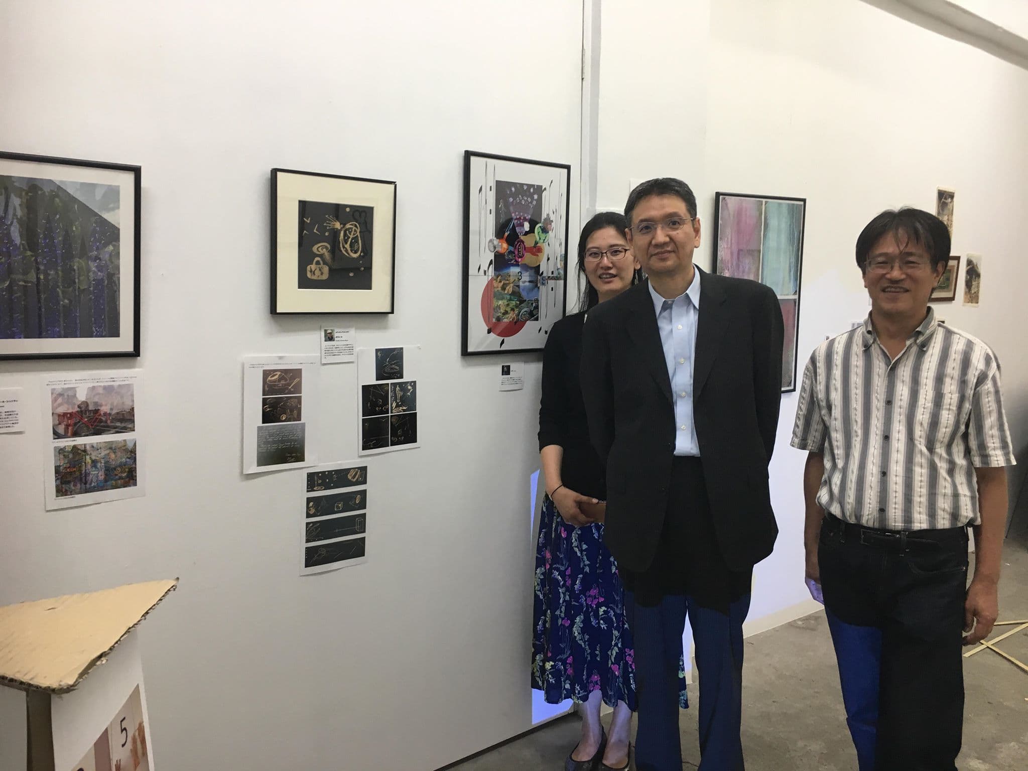 Three people posing in front of the hung wall, with framed works and small printed cards arranged around them