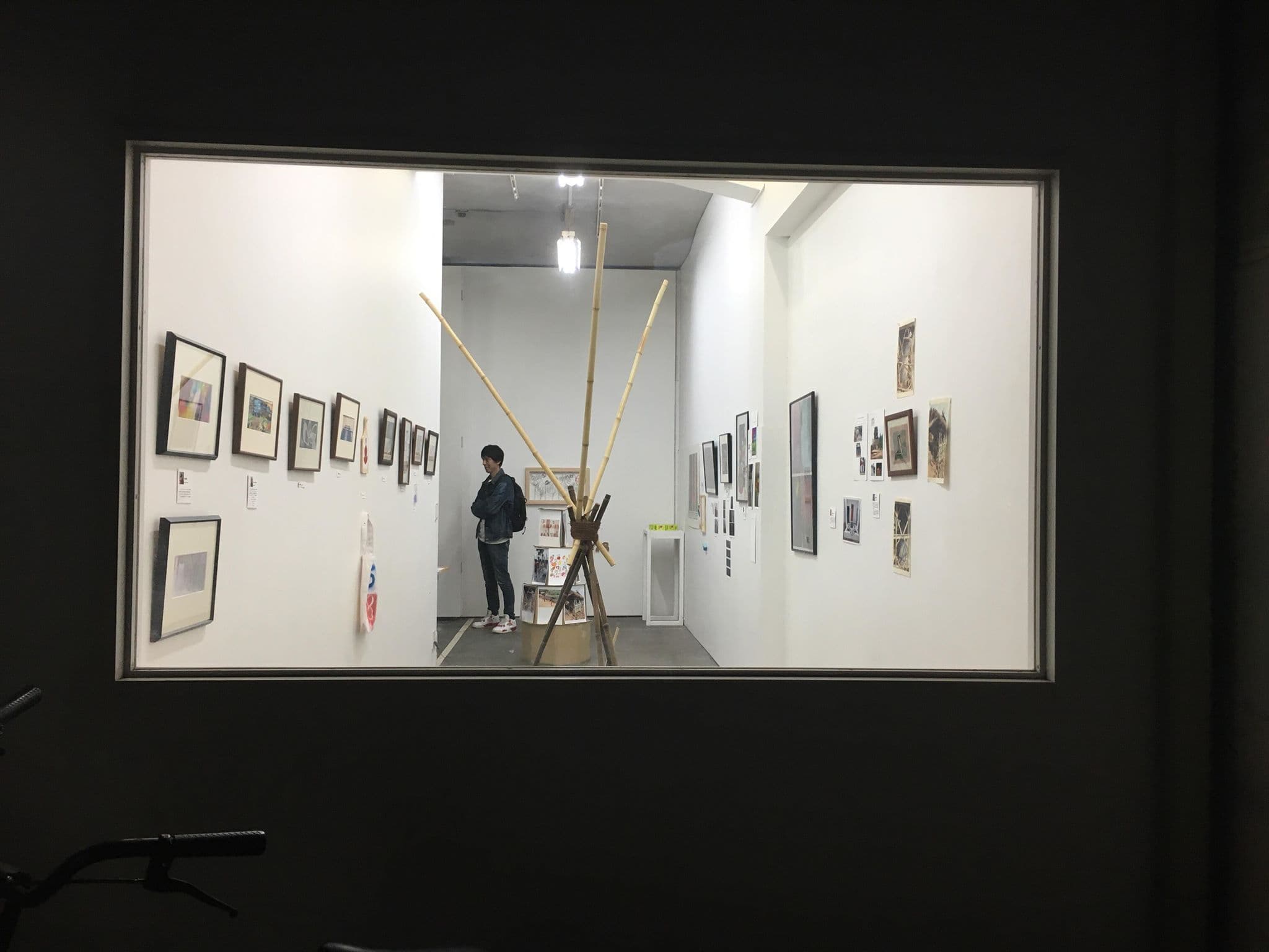The gallery seen through a window: framed works along both walls and a standing sculpture of crossed bamboo poles in the middle of the room