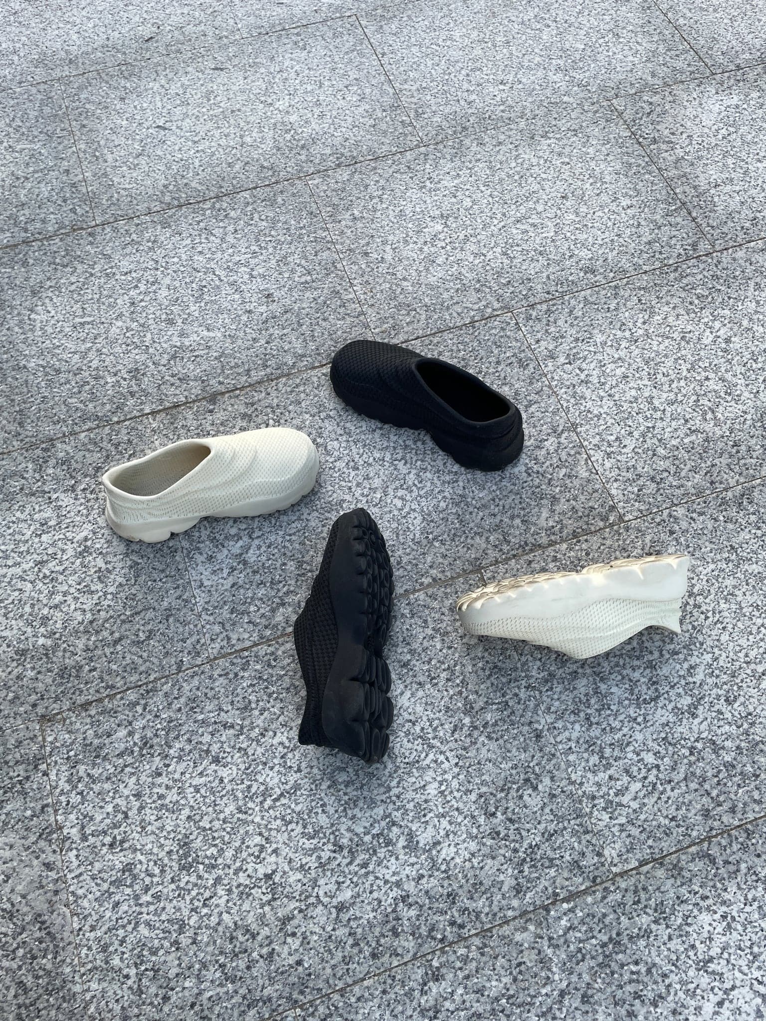 Four shoes arranged on pale granite paving: two bone and two black, one turned over to show the oval-pod tread.