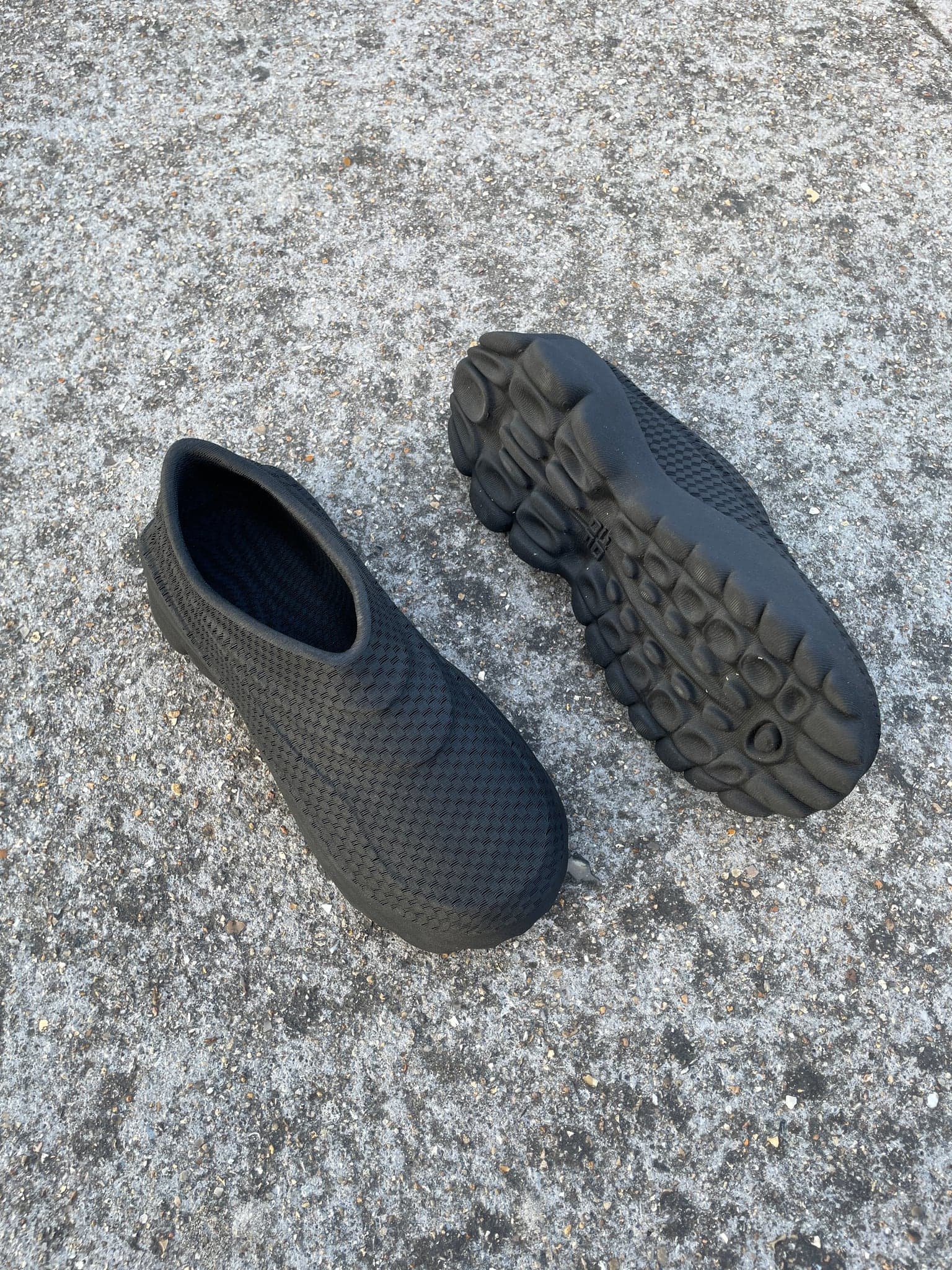 A black pair on rough concrete, one shoe upside down showing the pod tread, the other upright showing the knitted upper and open ankle.