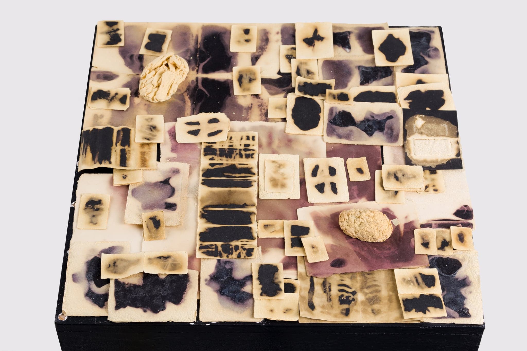 A low black square plinth photographed on white, its top surface tiled edge to edge with overlapping cream and violet wax-and-paper squares marked with black shapes.