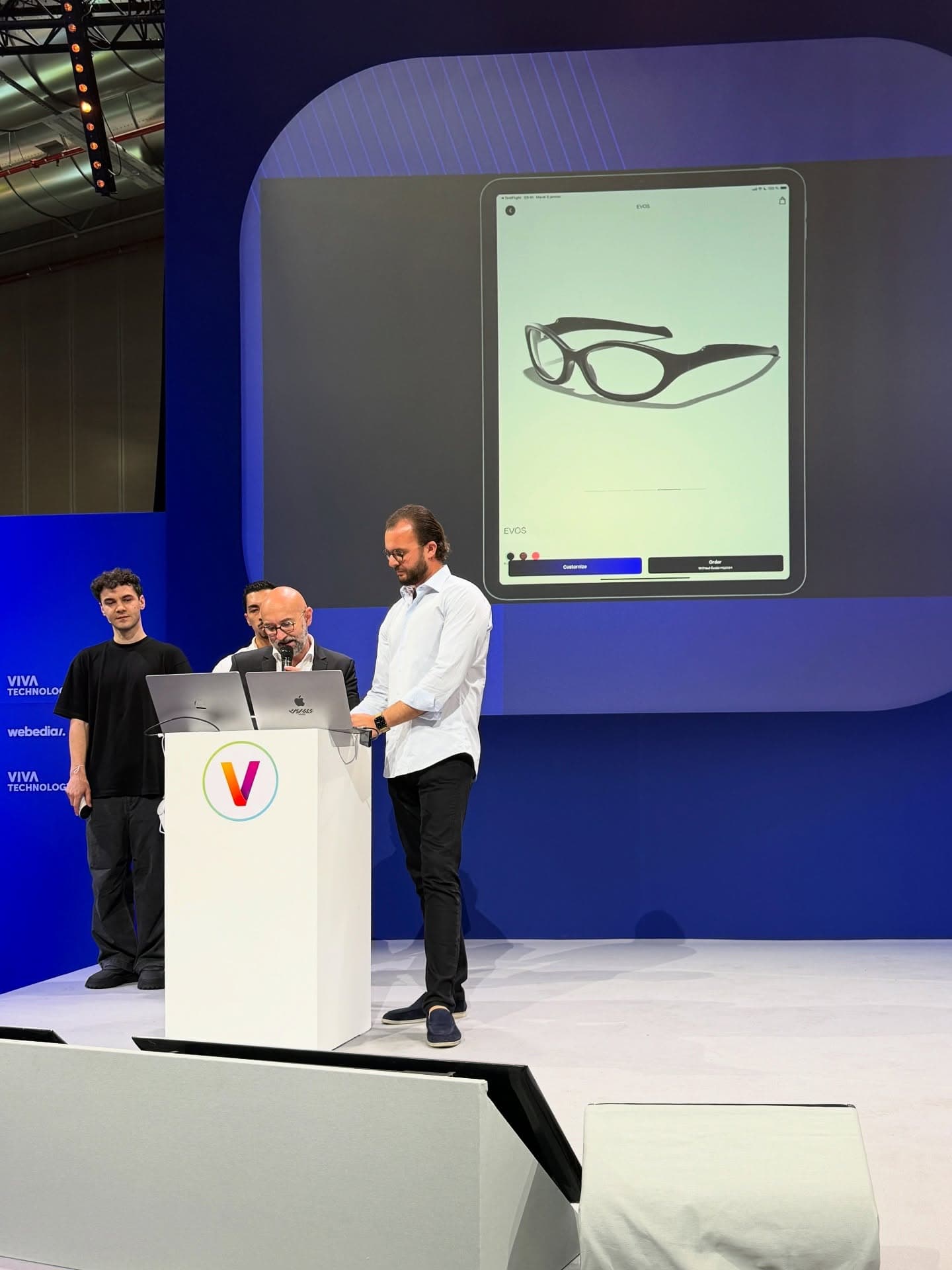Two people presenting on stage at VivaTech beside a lectern, the eyewear on the screen behind