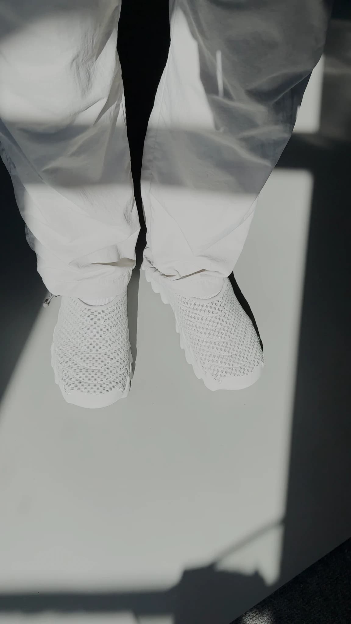 Looking straight down at both feet wearing VIZE with wide white trousers, the shoes reading almost as part of the floor