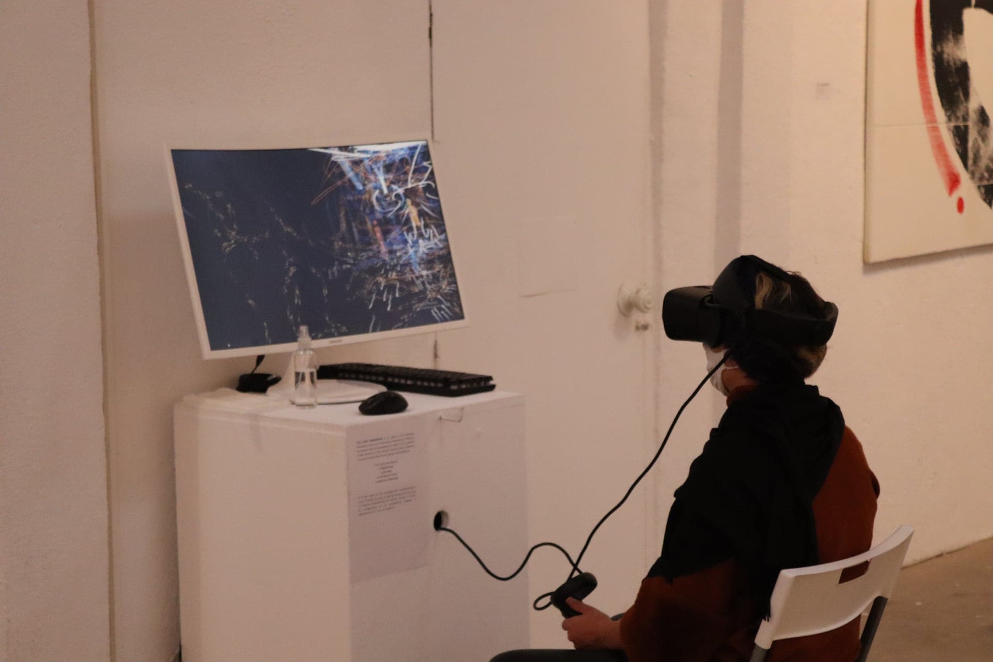 A visitor in a VR headset seated at a white plinth, the monitor beside them showing pale drawn strokes suspended in a dark space